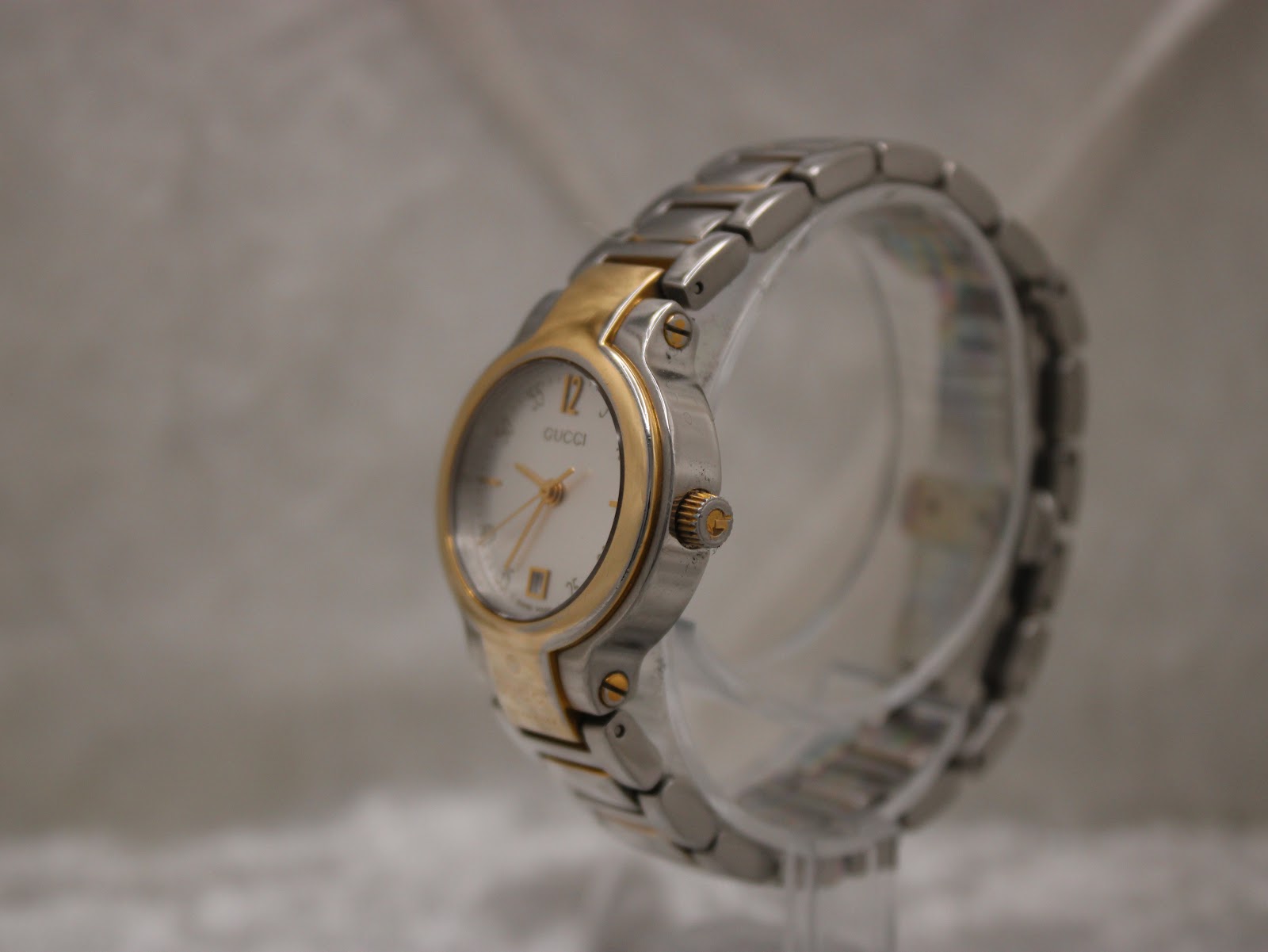 Angled side view of the Gucci 8900L watch on a clear display stand, highlighting the gold-tone crown and bracelet links.
