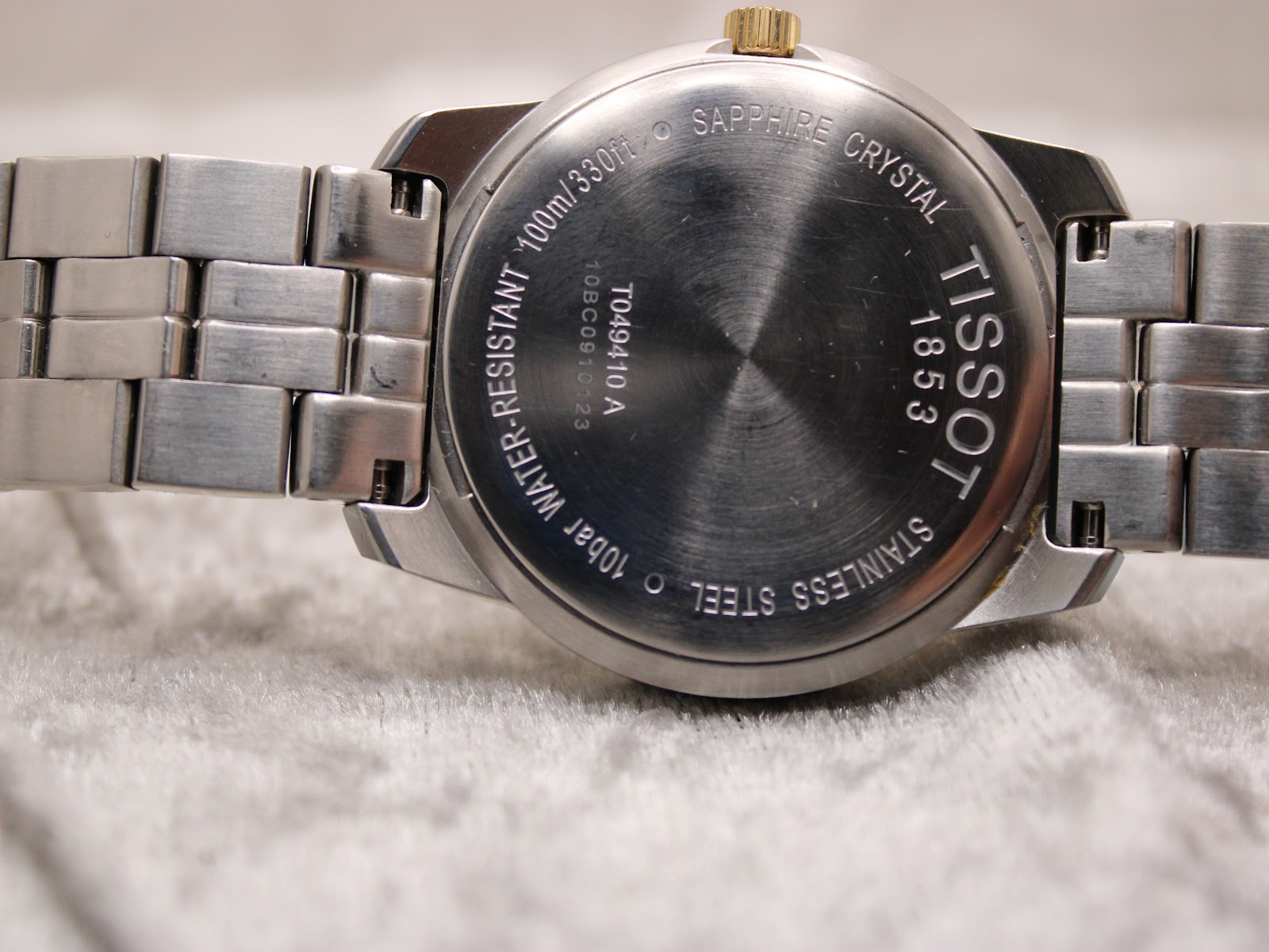 Close-up of the Tissot PR 100 watch case back showing 'Tissot 1853', 'Sapphire Crystal', '100m/330ft', 'Water-Resistant', 'Stainless Steel' and reference T049.410.A.