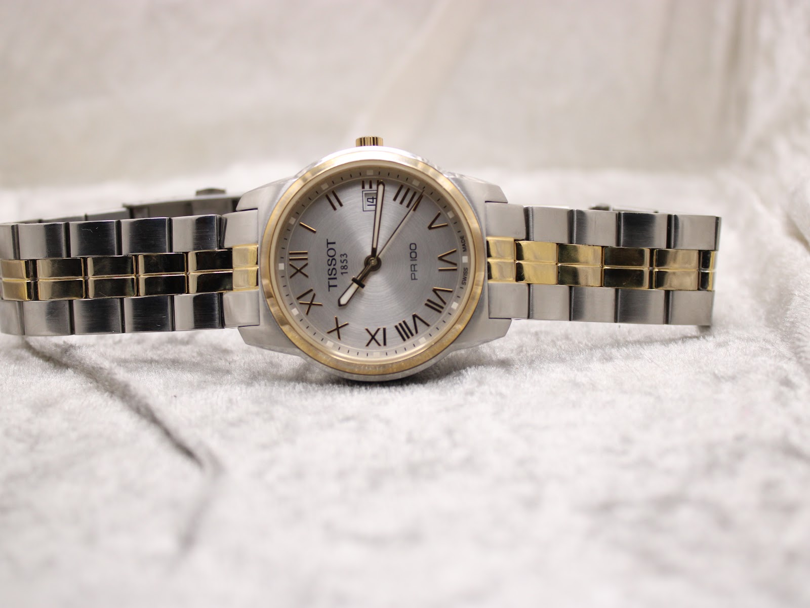 Front view of the Tissot PR 100 watch with its two-tone bracelet extended, on a soft white surface.