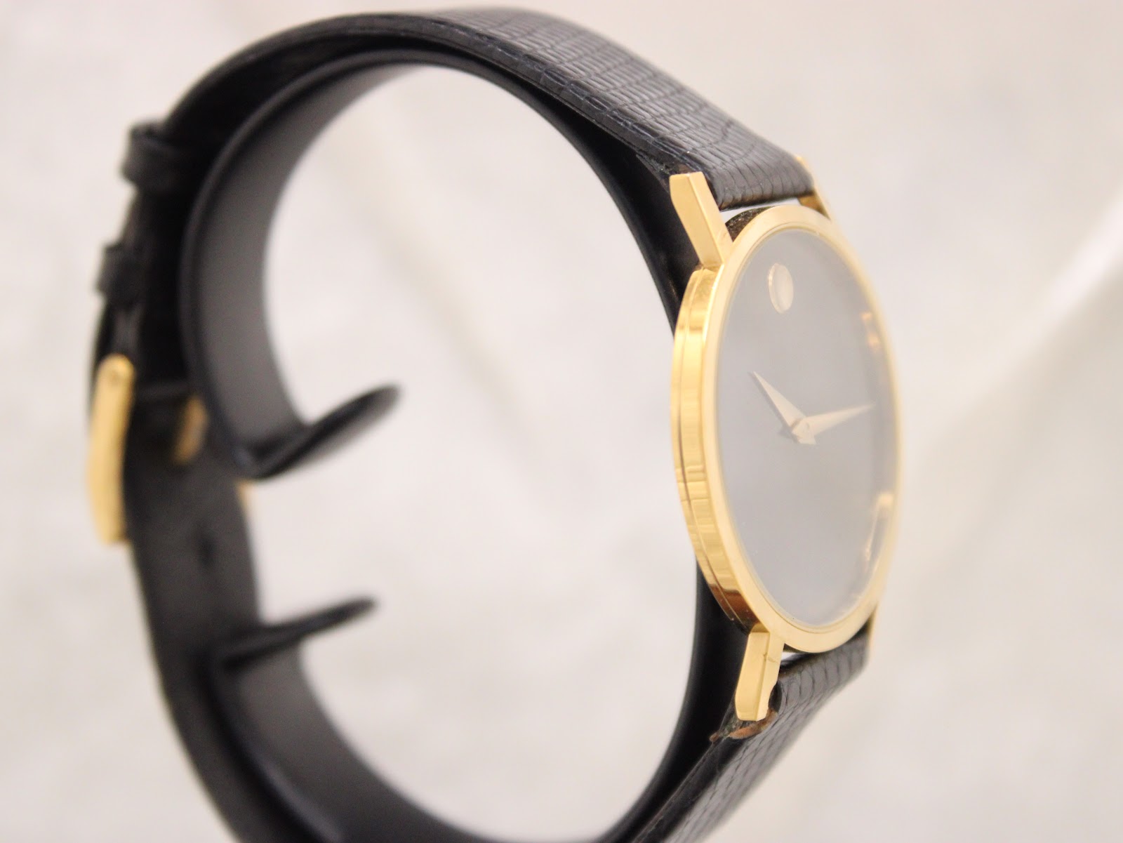 Angled side view of the Movado Museum watch, showing the gold-plated case and black leather strap.