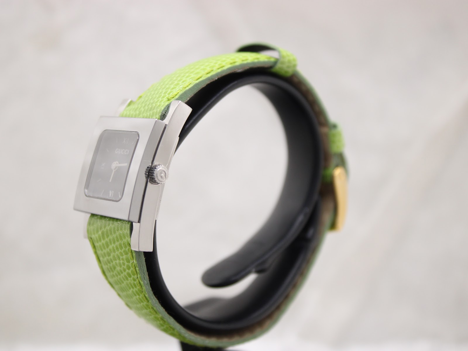 Side profile view of the Gucci 7900P watch, highlighting the stainless steel case and signed crown, with the green leather strap curving around.