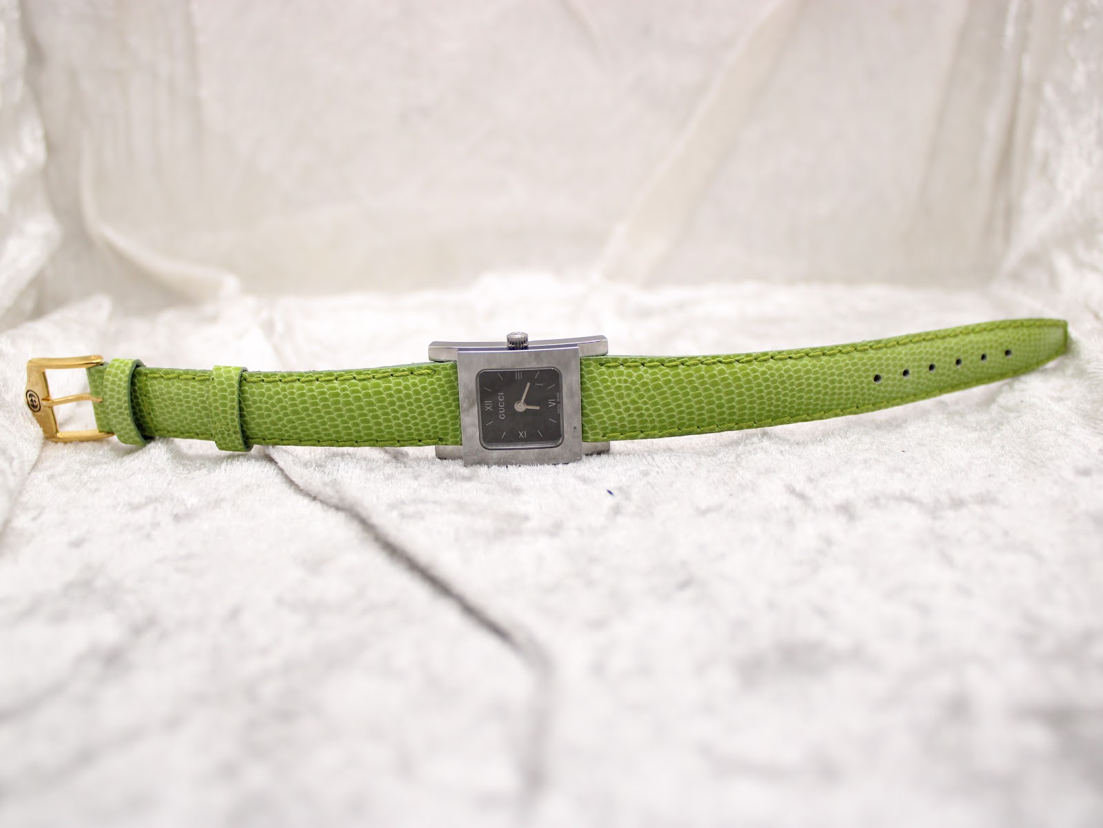 Full view of the Gucci 7900P watch laid flat on a white textured surface, showcasing the rectangular case and the long green leather strap.