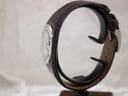 Side view of the Gucci 104 watch on a stand, highlighting the curved profile of the dark brown leather strap and stainless steel case.