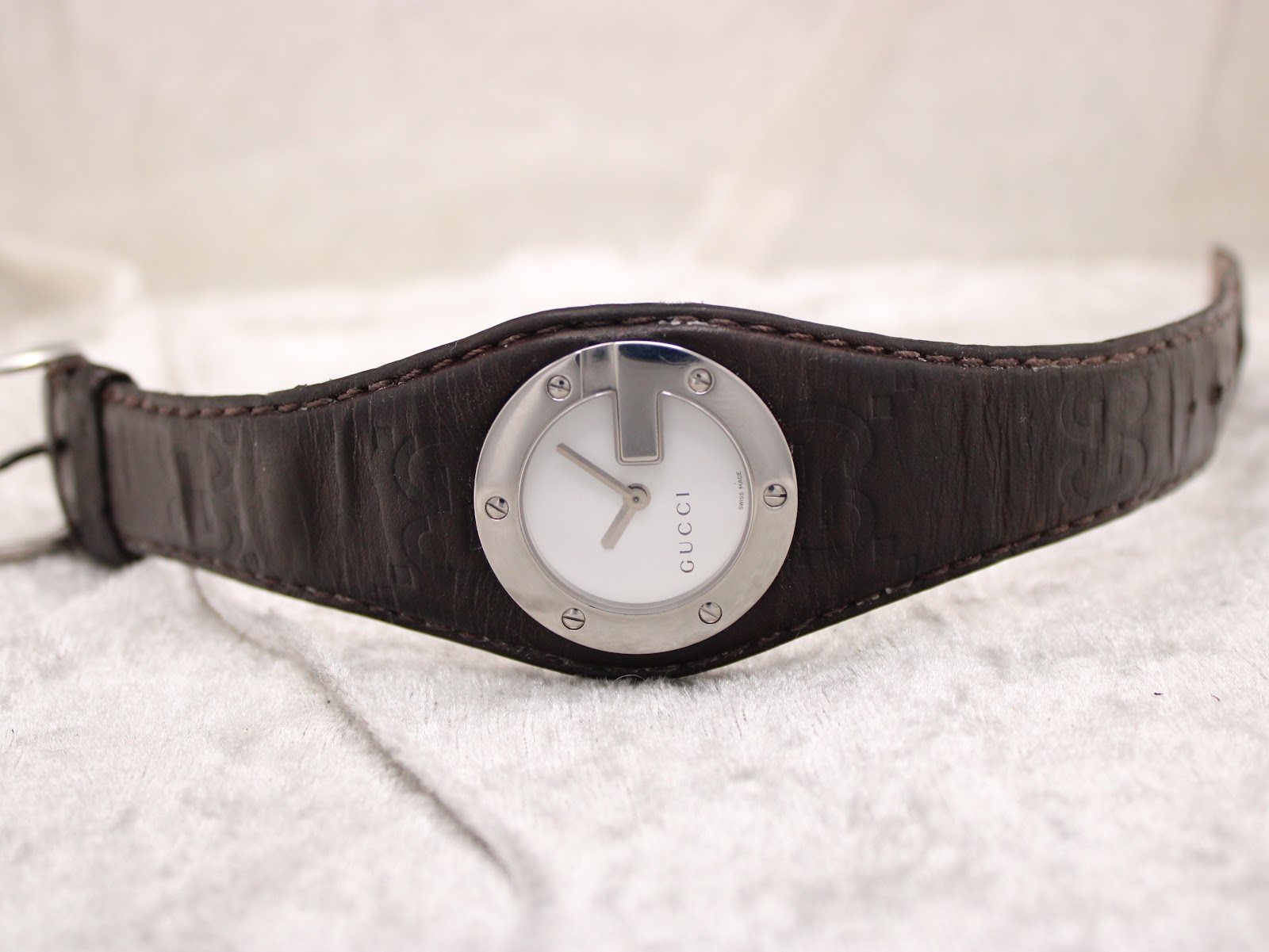 Top-down view of the Gucci 104 watch with its dark brown leather strap laid flat, showcasing the white dial and silver G-design case.
