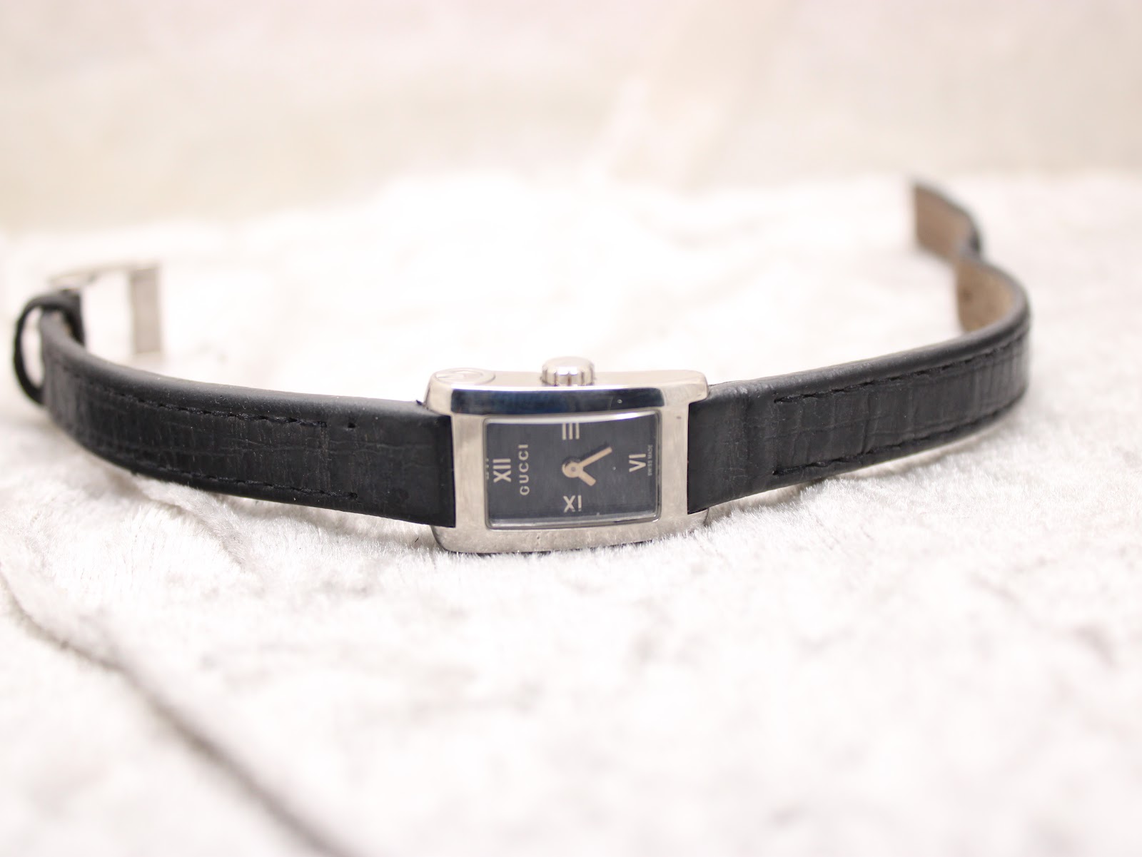 Top-down view of the Gucci 8600L watch laid flat on a textured white surface, displaying the black dial and black leather strap.
