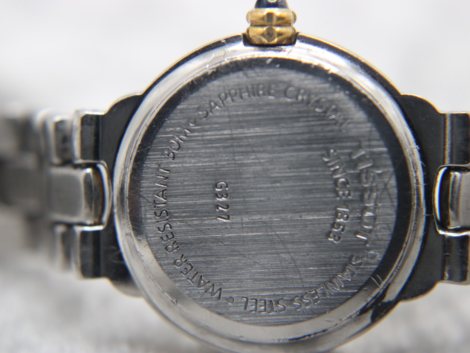 Close-up of Tissot G327 white dial with gold baton indices and blue cabochon crown detail