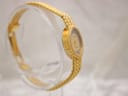 Angled view of the Tissot ladies' watch on a clear display stand, highlighting the woven gold-tone bracelet and oval dial.