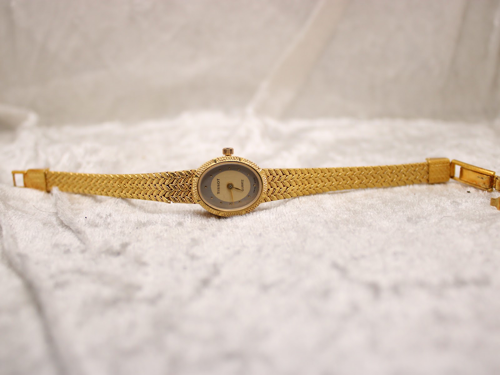 Top-down view of the Tissot gold-tone watch with its original woven bracelet unclasped and laid flat on a textured white surface.