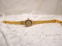 Top-down view of the Tissot gold-tone watch with its original woven bracelet unclasped and laid flat on a textured white surface.