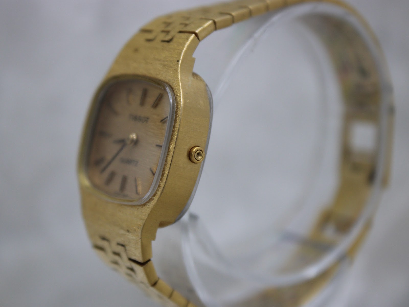Side view of the Tissot watch on a clear display stand, highlighting the brushed gold case and recessed crown.