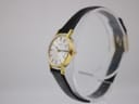 Angled side view of the Tissot Stylist watch showing the gold-plated case, crown, and black strap on a stand.