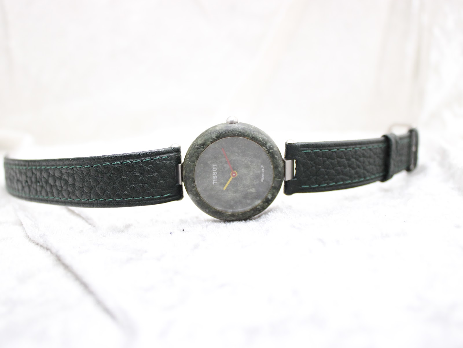 Full front view of the Tissot RockWatch and its green leather strap laid flat on a white textured surface.