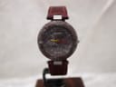 Front view of Tissot RockWatch with burgundy natural stone dial, minimalist design, yellow and red hands on a stand.