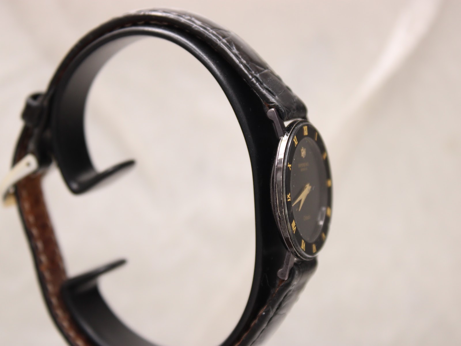 Three-quarter view of the Raymond Weil Othello watch, showing the left side of the case and the black leather strap on a watch stand.