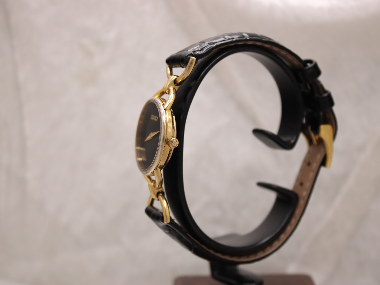 Side profile view of the Gucci watch on a stand, highlighting the gold-tone case and crown against the black leather strap.