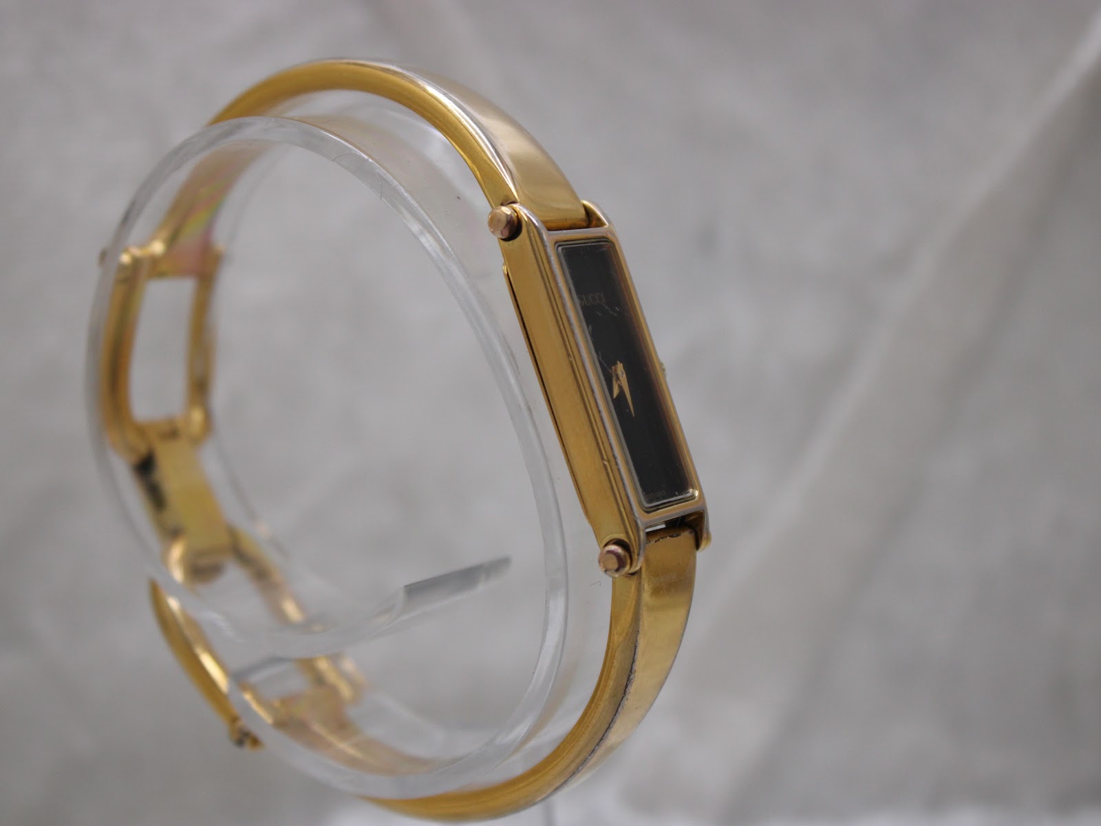 Another angled side view of the gold plated Gucci 1500L watch on a clear display stand, showing the full bangle shape.