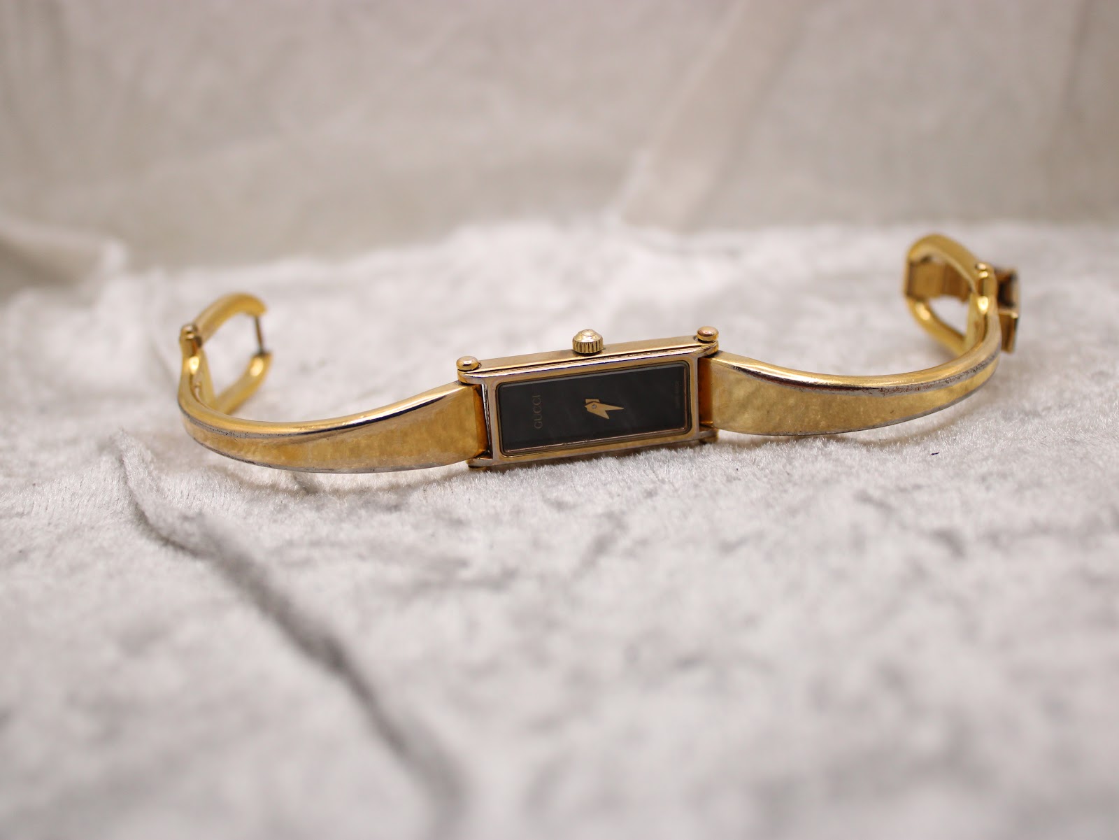 Top-down view of the gold plated Gucci 1500L bangle watch with clasp open, laid flat on a white textured surface.