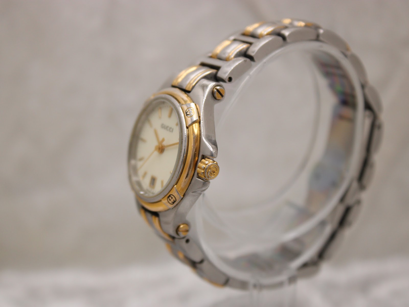 Angled side view of the Gucci 9040L watch showing the gold-tone crown and case profile on a clear stand.