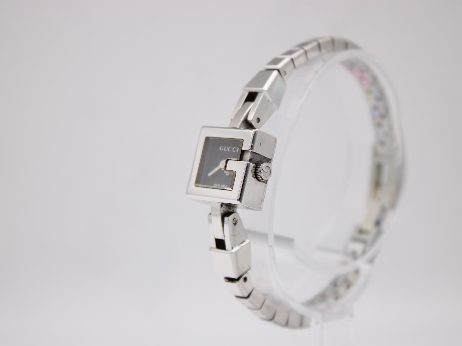Angled side view of the Gucci G-Frame watch showing the case side and original signed crown, on a clear stand.