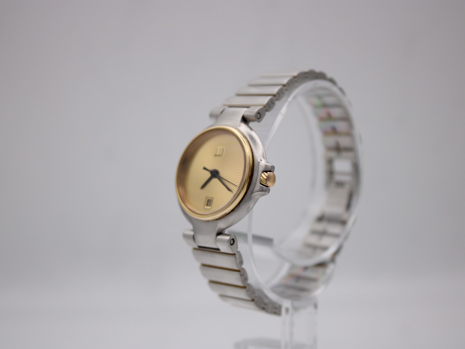 Angled side view of the Dunhill watch on a clear stand, showing gold bezel and two-tone bracelet.