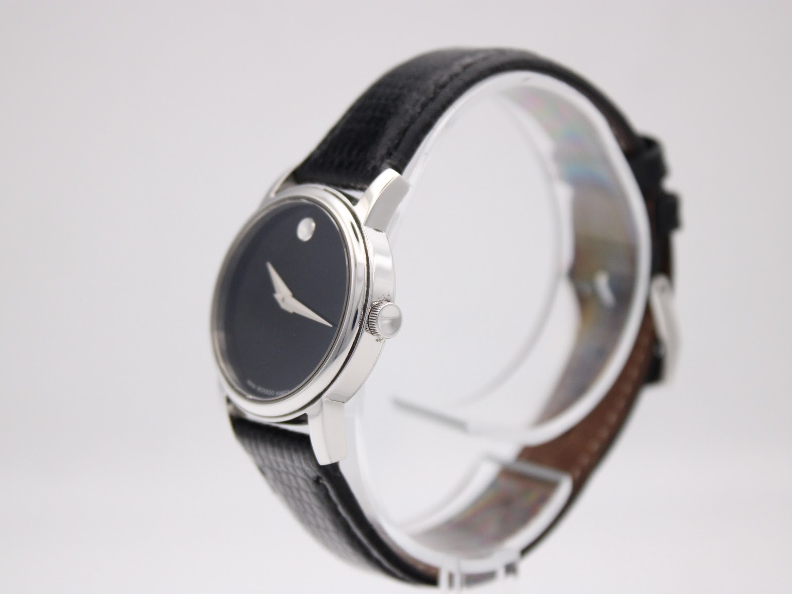 Angled side view of the Movado Museum watch on a clear display stand, showing the minimalist case profile and textured black leather strap.