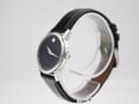 Angled side view of the Movado Museum watch on a clear display stand, showing the minimalist case profile and textured black leather strap.