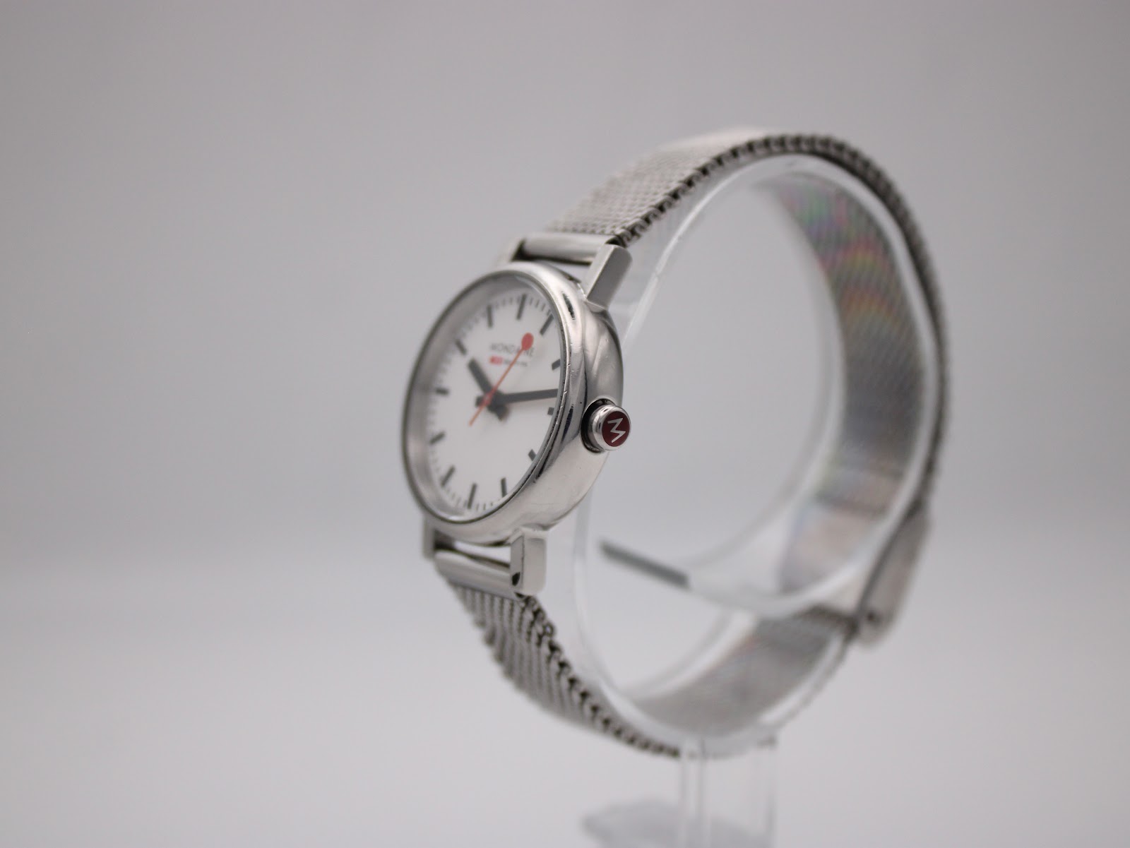 Side angled view of Mondaine watch showing stainless steel case and mesh bracelet on a display stand.