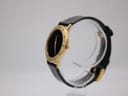 Angled side view of the Gucci 3000.2.M watch, showcasing its slim gold-tone case and black leather strap on a clear display stand.