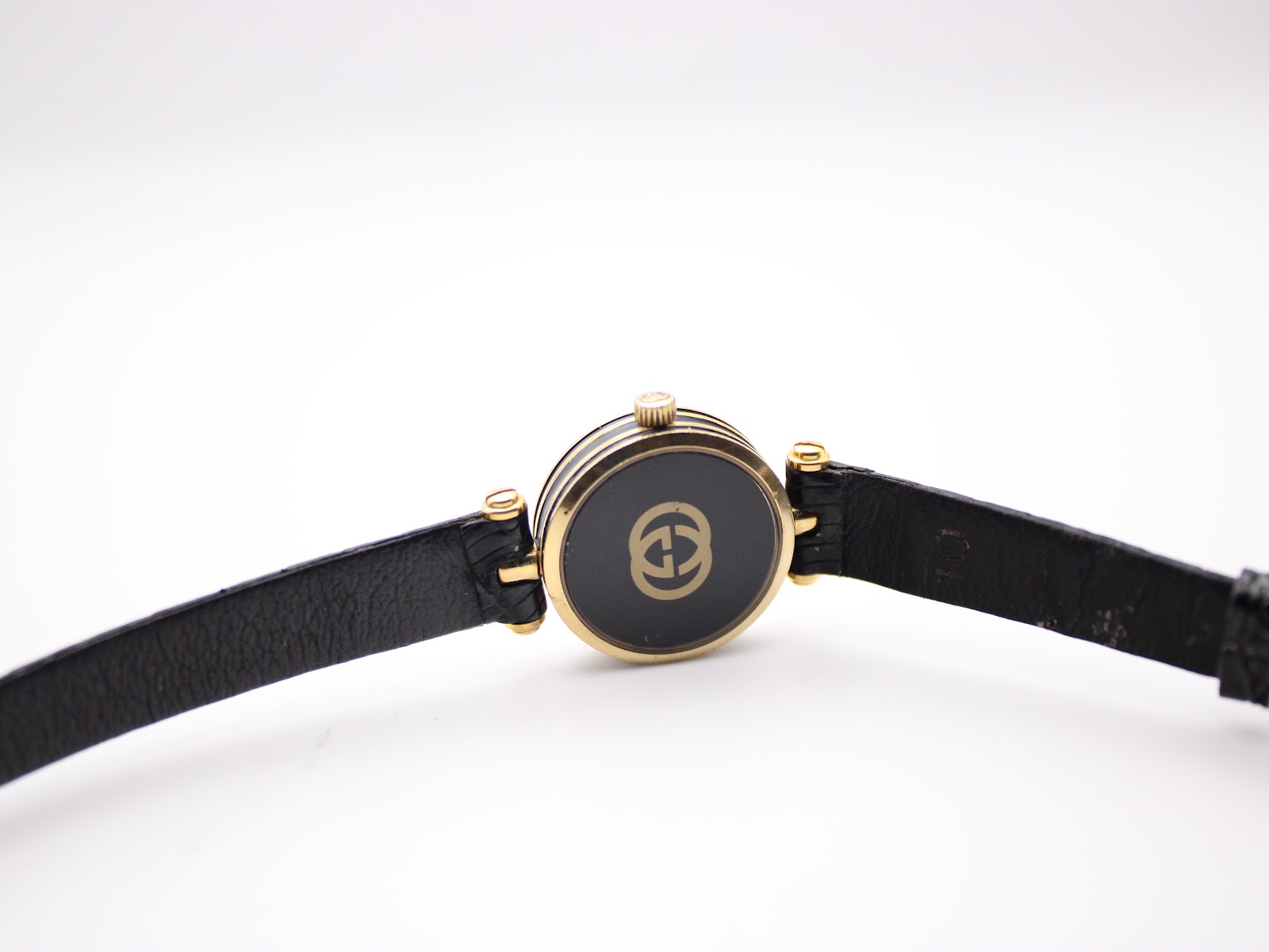 View of the watch from the back, showing the gold-plated case back with the Gucci logo and the black leather strap extended.