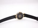 View of the watch from the back, showing the gold-plated case back with the Gucci logo and the black leather strap extended.