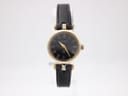 Front view of the Gucci 2000L watch, showing the black dial with gold Roman numerals and gold-plated case, on a clear stand.