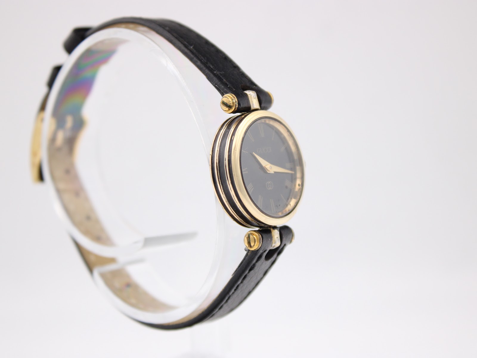 Angled side view of the Gucci 2000L watch, showing the black dial and gold case detail on a clear stand.