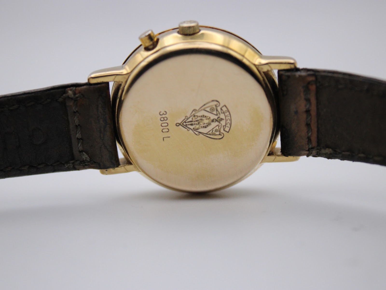 Close-up of the watch case back, showing the embossed Gucci crest, '3800 L', and serial number with part of the brown leather strap.