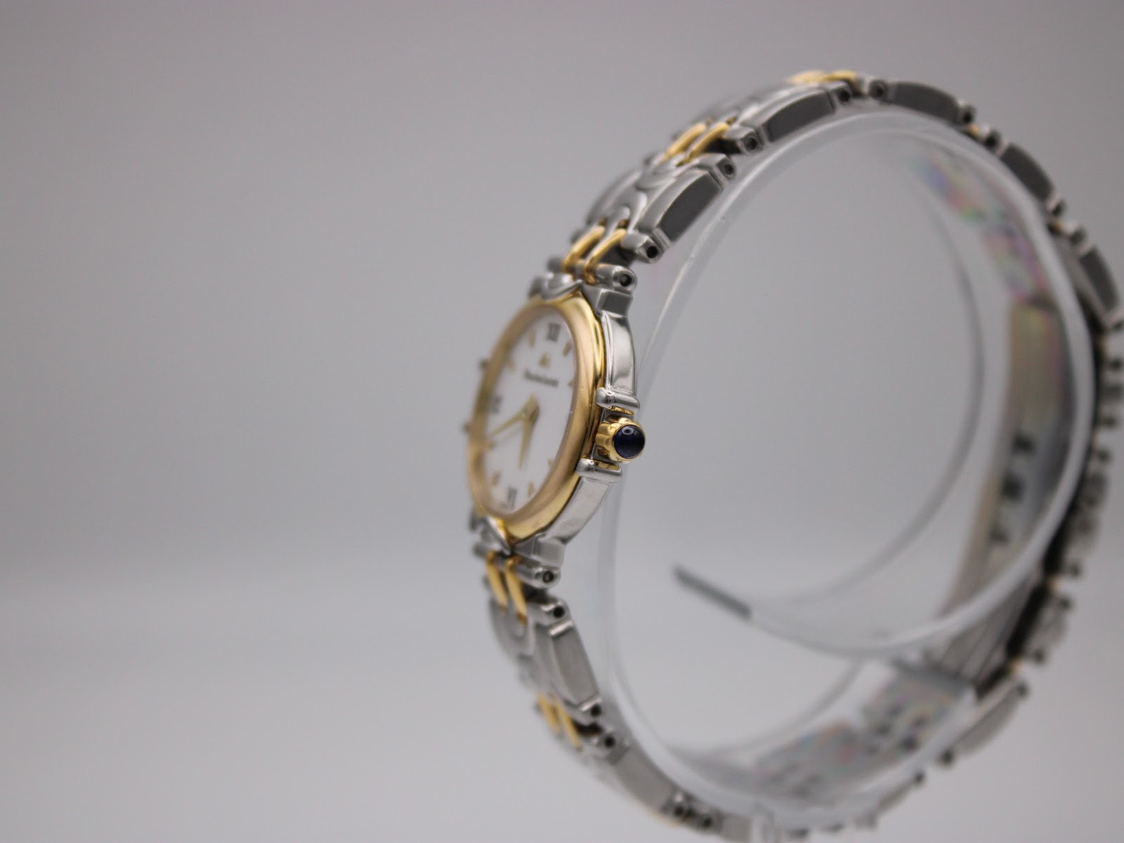 Side profile of the Maurice Lacroix watch on a clear stand, highlighting the gold-tone crown with a blue cabochon.