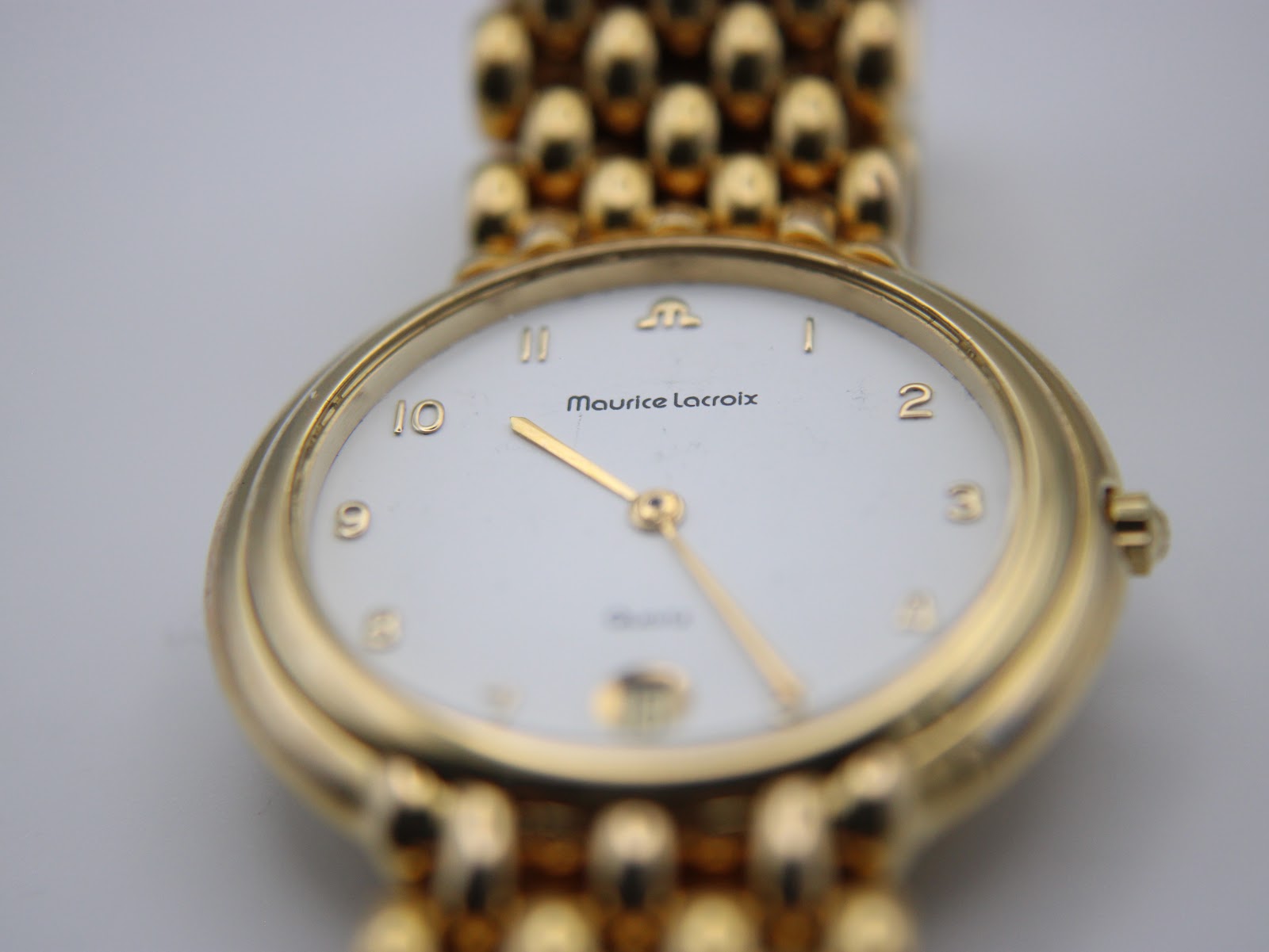 Angled close-up of the Maurice Lacroix watch dial, showing the white face, gold Arabic numerals, date window at 6, and 'Maurice Lacroix Quartz' text.