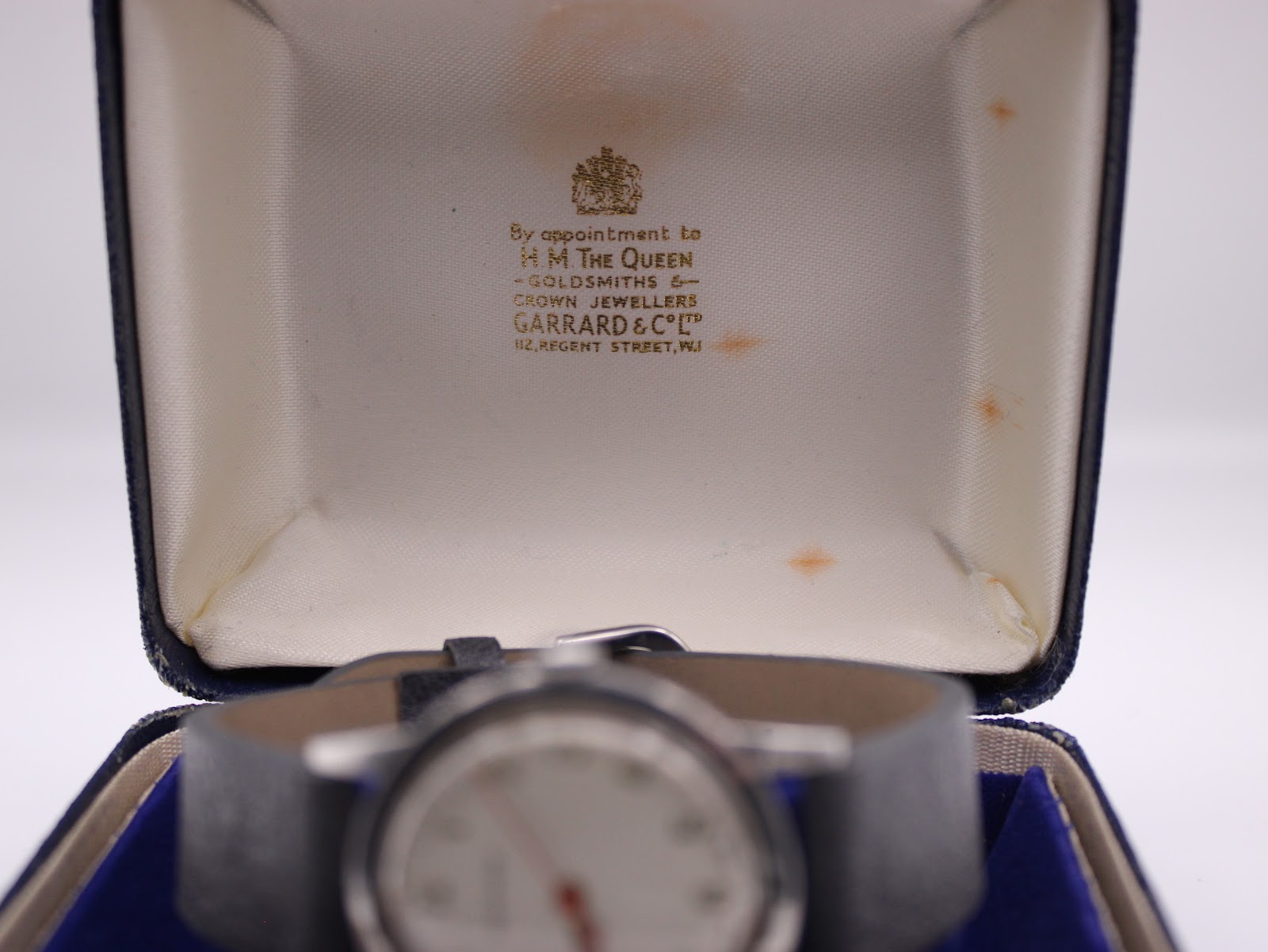 Interior of the original Garrard presentation box, lined with cream silk, showing 'By appointment to H.M. THE QUEEN' text, with the watch visible but blurred in the foreground.