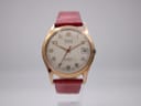 Front view of the Rone automatic watch, showing the gold-tone case, cream dial with Arabic numerals, red seconds hand, and date window, on a red leather strap.