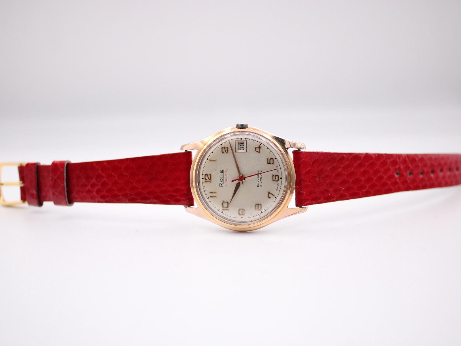 Full view of the Rone watch laid flat, displaying the red leather strap with gold-tone buckle and the cream dial.