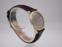 Another angled side view of the watch on a stand, showing the curved brown leather strap and the slim profile of the gold case.