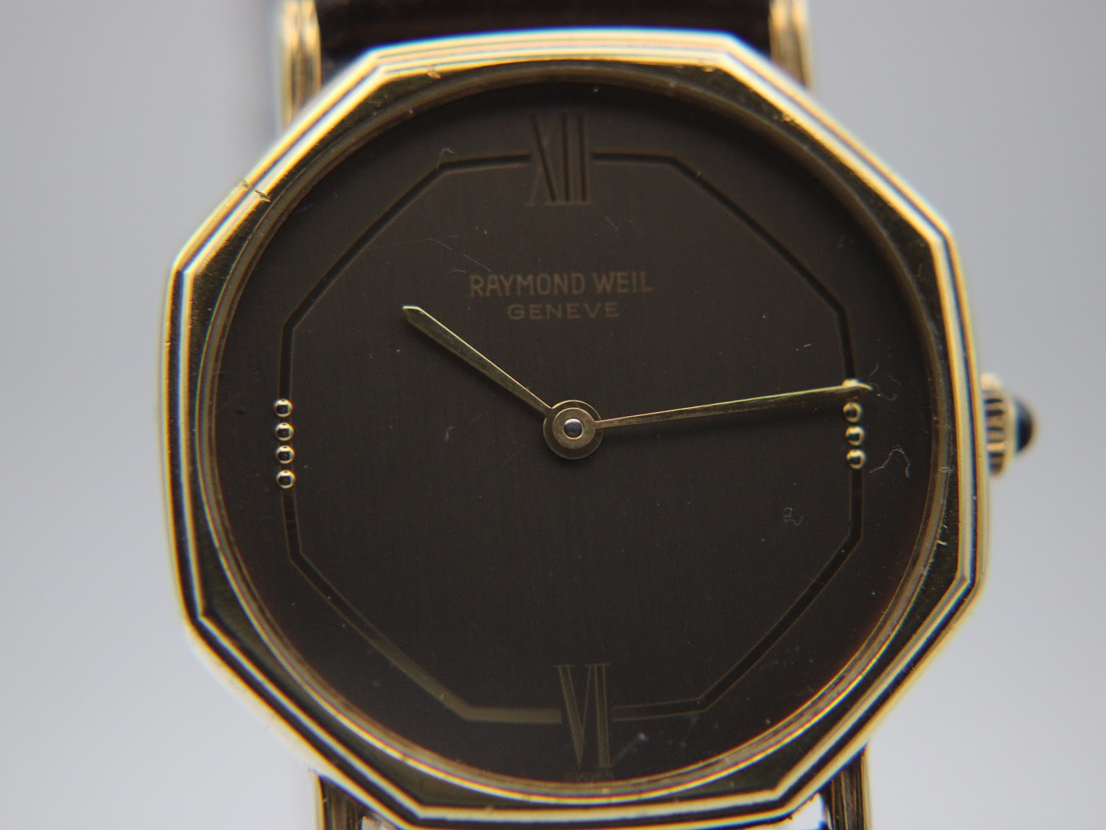 Close-up front view of the Raymond Weil Genève watch dial, showing the gilt baton indices, Roman numerals at XII and VI, and minimalist two-hand layout.