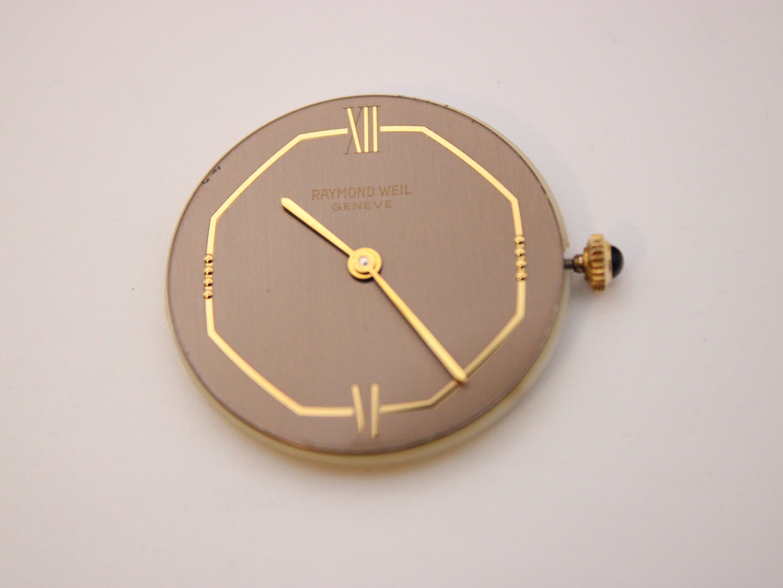 Close-up view of the Raymond Weil Genève brown dial and gold hands inside the octagonal case, with the black cabochon crown visible.