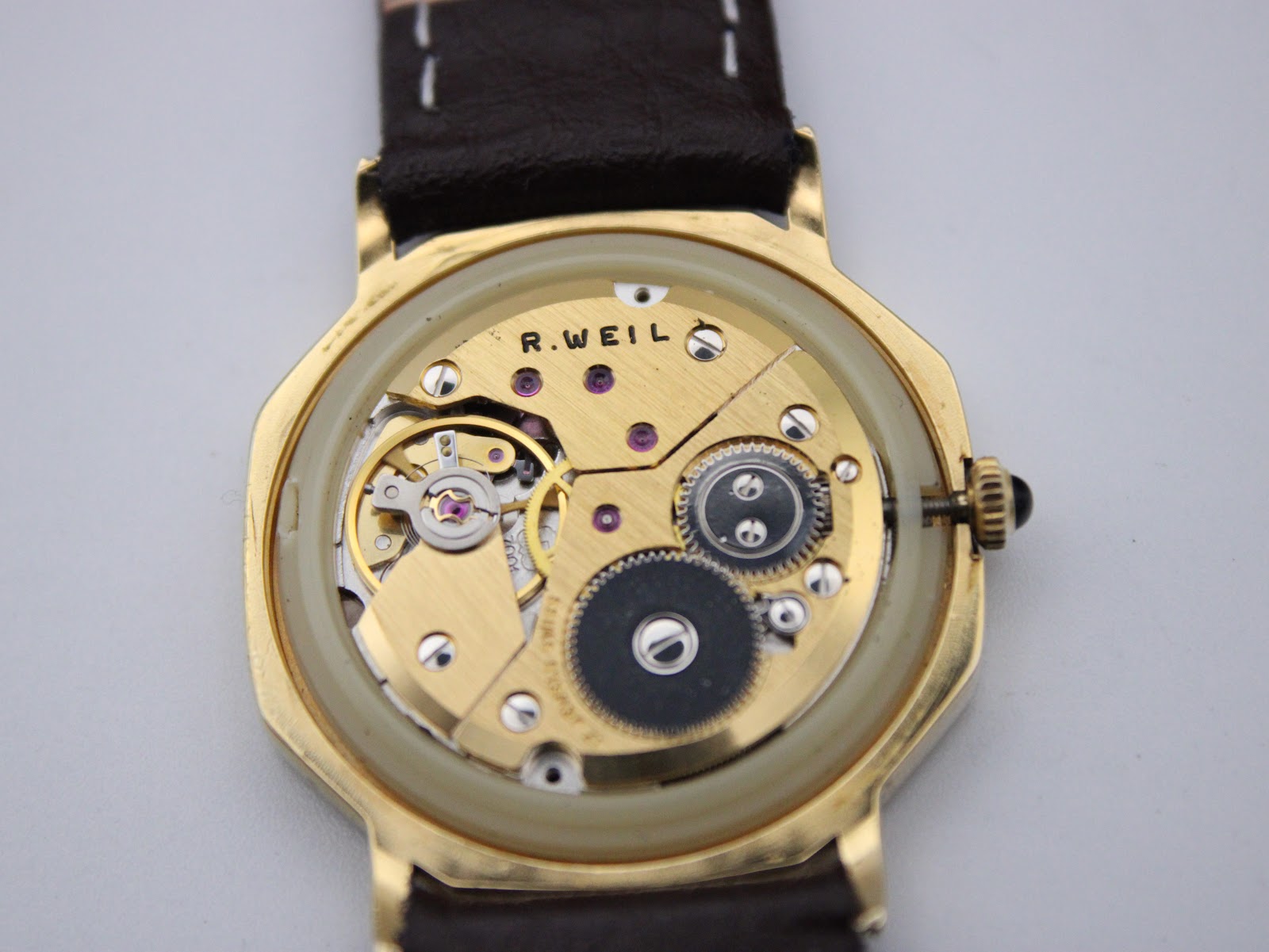 Close-up view of the manual-wind movement inside the gold-plated case, showing the "R. WEIL" engraving and intricate gears.
