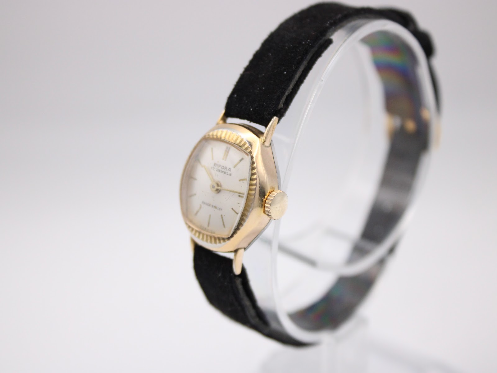 Angled view of the Bifora watch, showing the fluted bezel and gold-tone crown, on a clear stand.