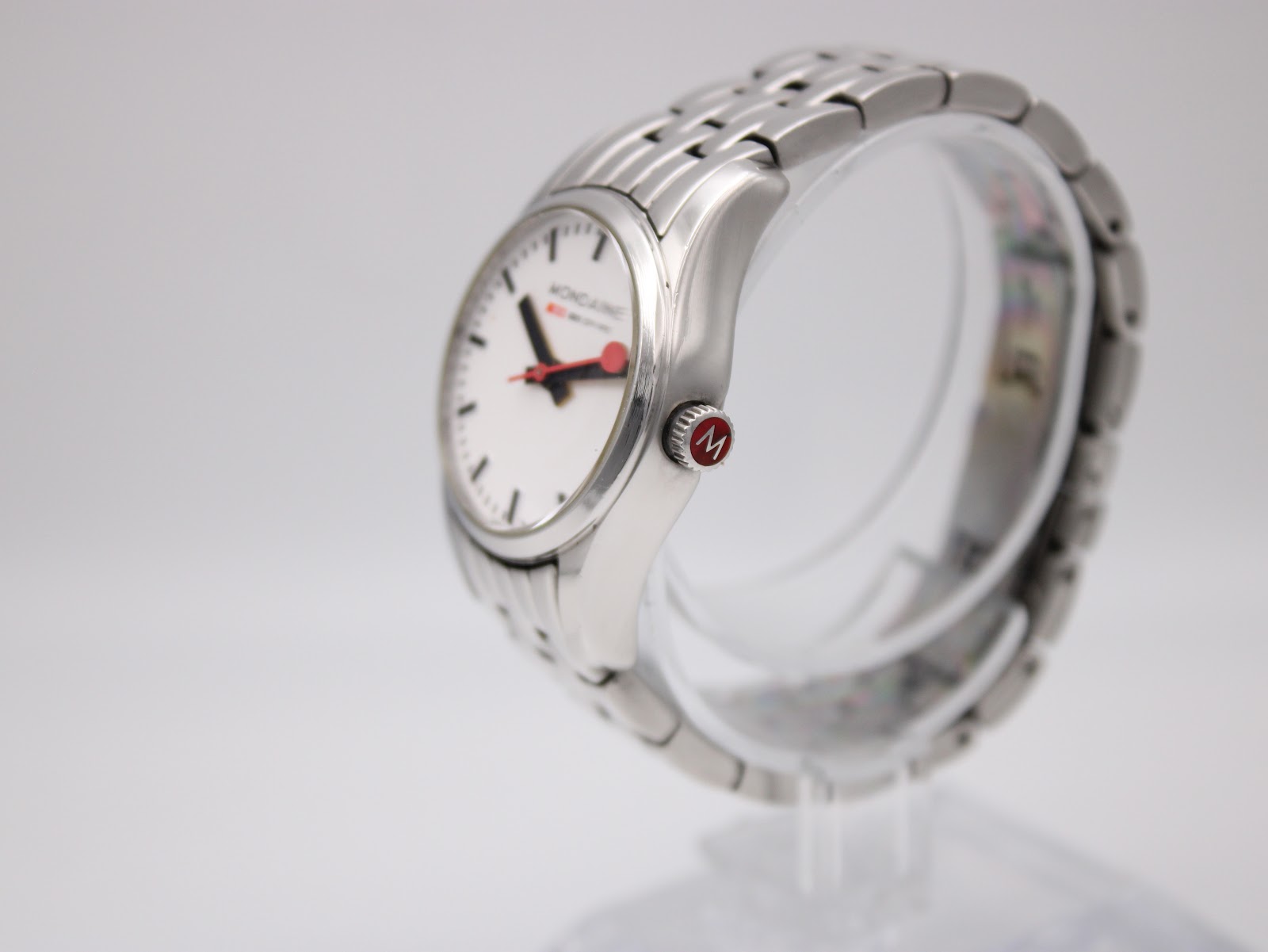 Side view of the Mondaine watch showing the polished stainless steel case and bracelet, with the red-detailed crown visible.