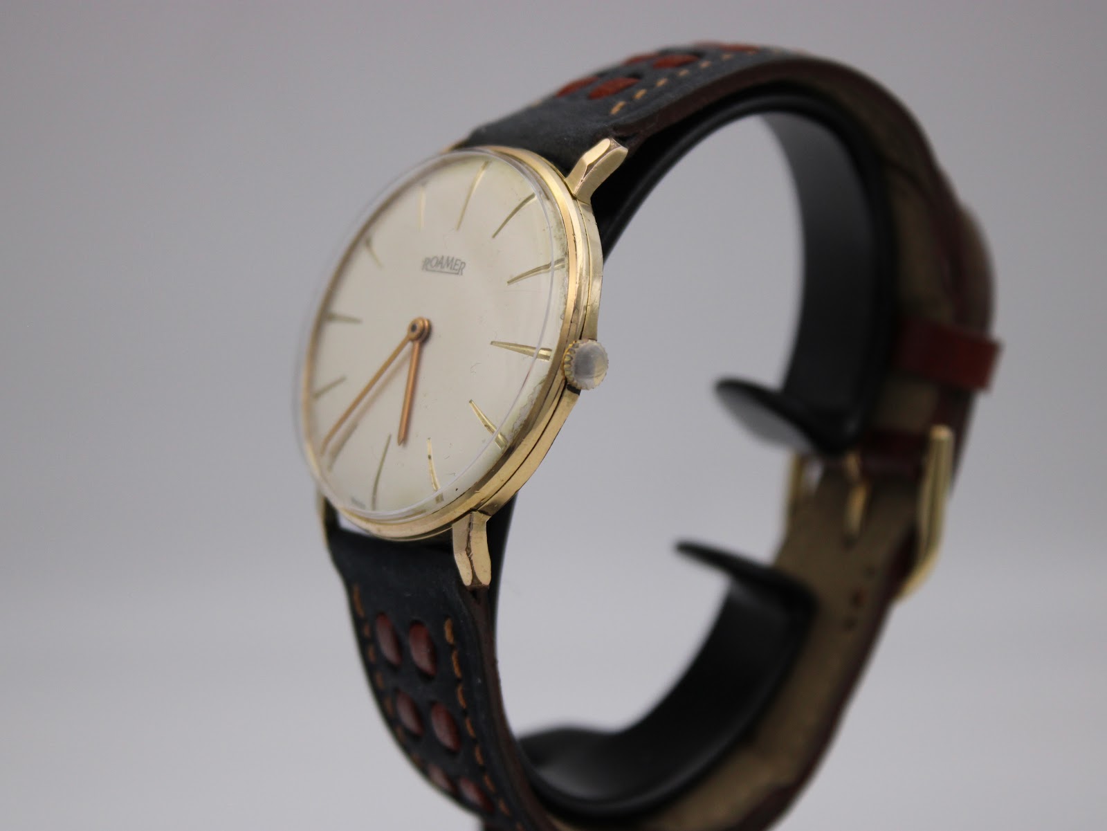 Side angled view of the gold plated Roamer watch on a stand, highlighting its slim case profile and domed acrylic crystal.