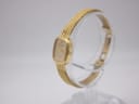 Angled view of the gold Raymond Weil 5707 watch on a clear stand, showing the rectangular case and crown.