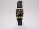 Front view of gold tone Bulova SL200 watch on a stand, showing black dial with gold baton markers and hands, and black leather strap.