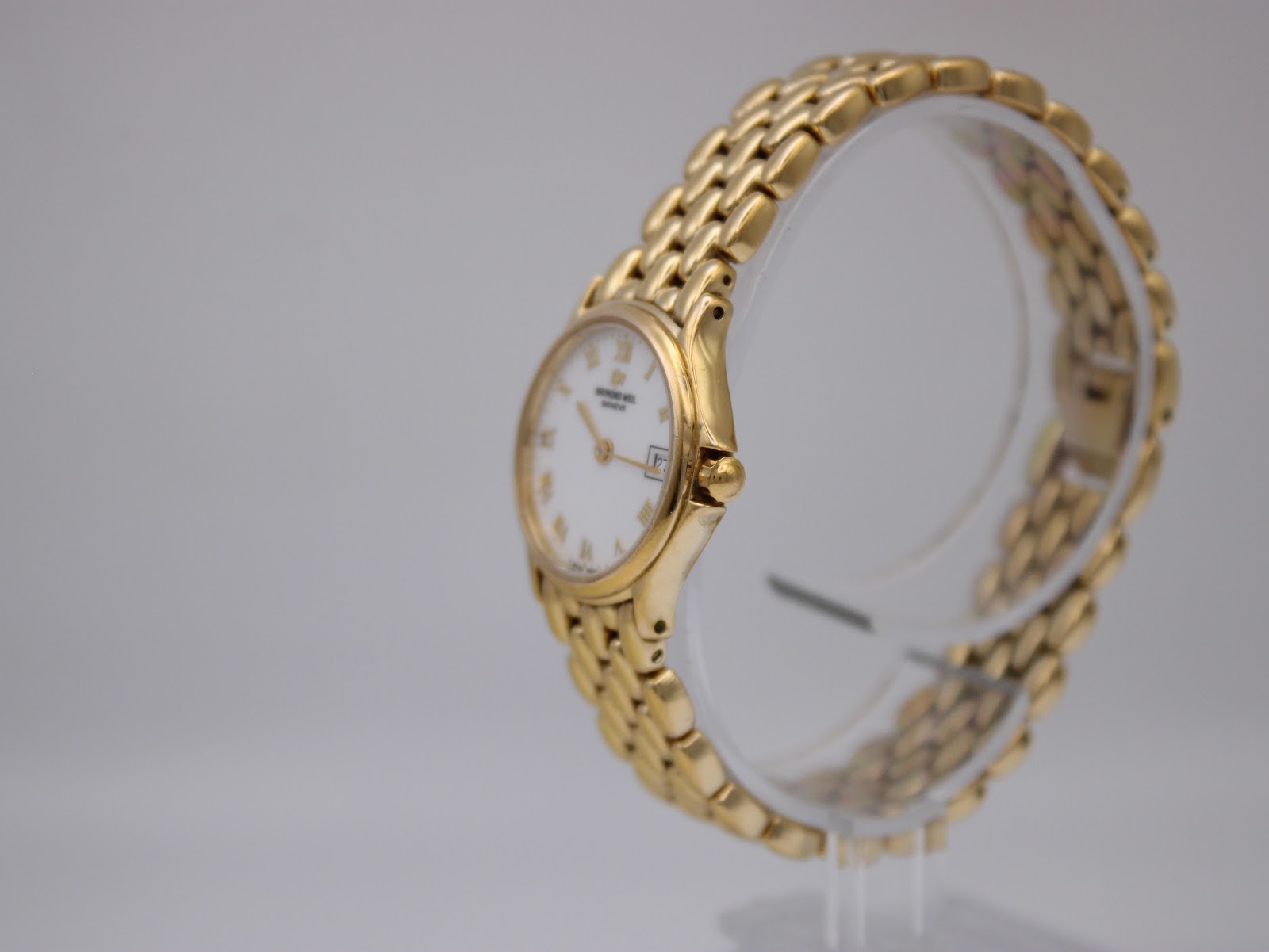Oblique side view of the gold Raymond Weil watch and bracelet on a clear display stand, highlighting the crown.