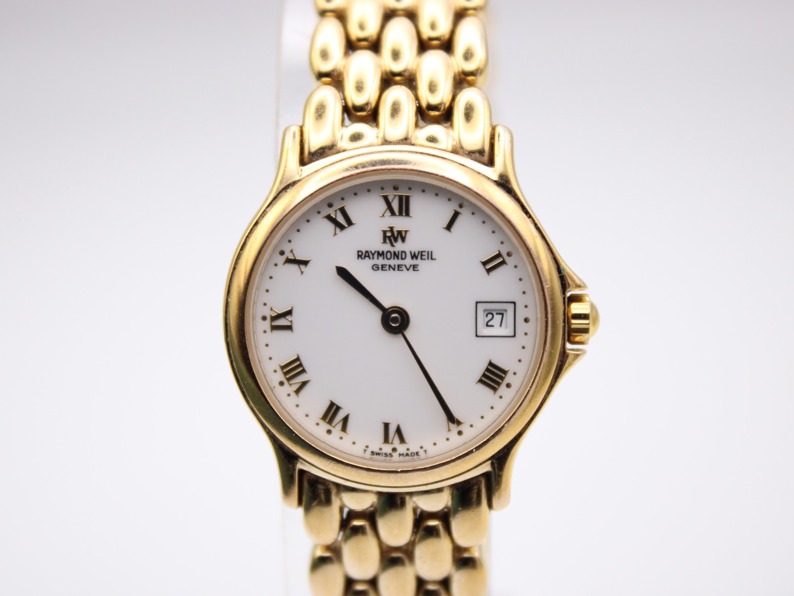Close-up front view of the Raymond Weil Genève white dial with prominent gold Roman numerals, a date window at 3 o'clock, and gold tone hands.