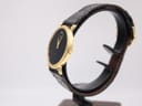 Angled side view of the Raymond Weil watch showing its slim gold case and black leather strap displayed on a watch stand.